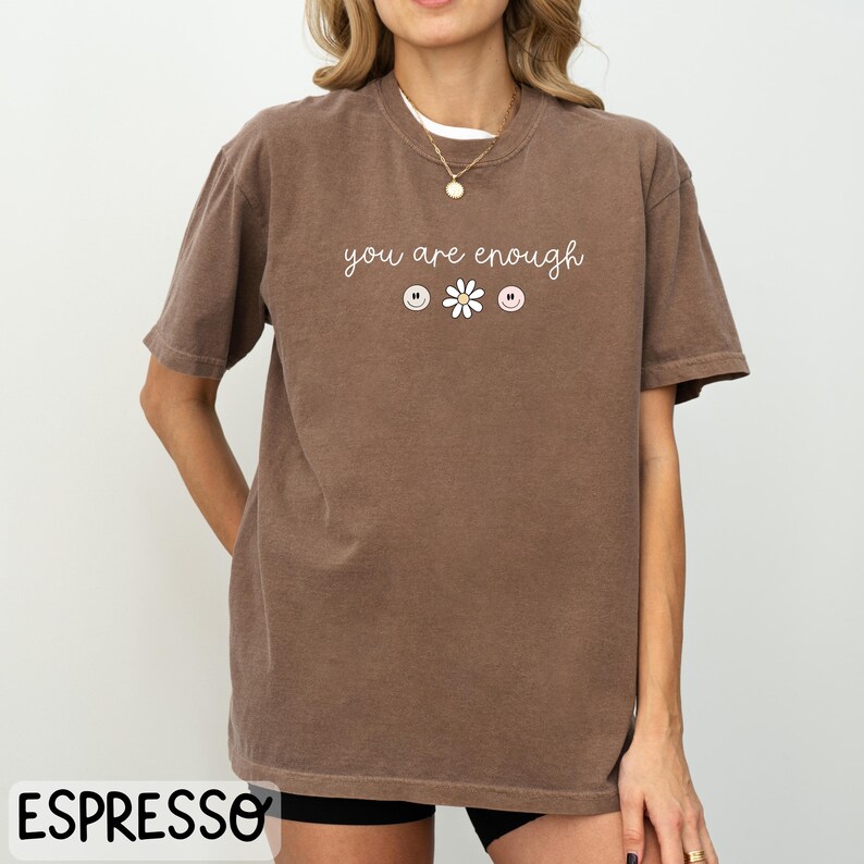 May include: A brown t-shirt with the text "you are enough" in white lettering. The shirt has three small graphic designs: two smiley faces and a daisy.