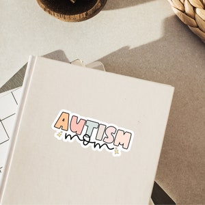 May include: A white sticker with the text "AUTISM mom" in rainbow colours with gold stars on a white notebook.