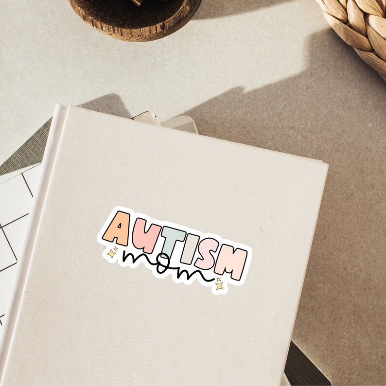 May include: A white sticker with the text "AUTISM mom" in rainbow colours with gold stars on a white notebook.