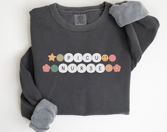 Puede incluir: Una sudadera gris oscuro con las palabras "PICU NURSE" escritas en círculos de colores. Cada letra está en un círculo de color diferente con un diseño de estrella, flor y cara sonriente.