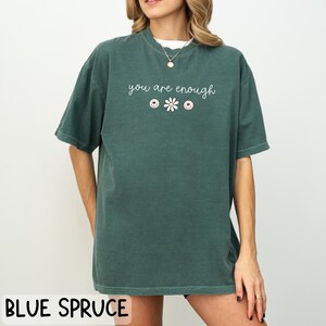 May include: A dark green t-shirt with the text "you are enough" in white lettering. The text is centred on the shirt and is accompanied by a small white daisy and three small pink and white buttons.