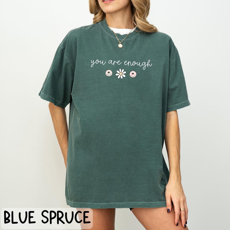 May include: A dark green t-shirt with the text "you are enough" in white lettering. The text is centred on the shirt and is accompanied by a small white daisy and three small pink and white buttons.