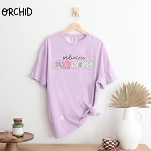 May include: A light purple t-shirt with the text "pediatric nurse" in colorful letters. The letters are decorated with hearts, stars, and other designs.