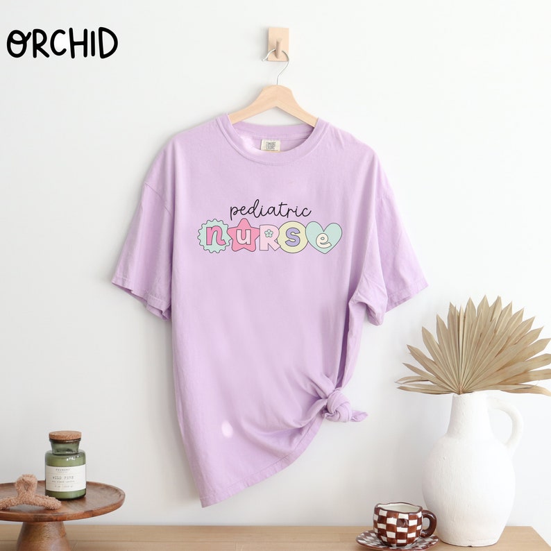May include: A light purple t-shirt with the text "pediatric nurse" in colorful letters. The letters are decorated with hearts, stars, and other designs.