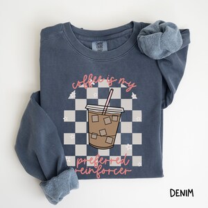 May include: Denim blue sweatshirt with a graphic of an iced coffee drink on a checkered background. The text "coffee is my preferred reinforcer" is in pink script. The sweatshirt has a crew neck and long sleeves.