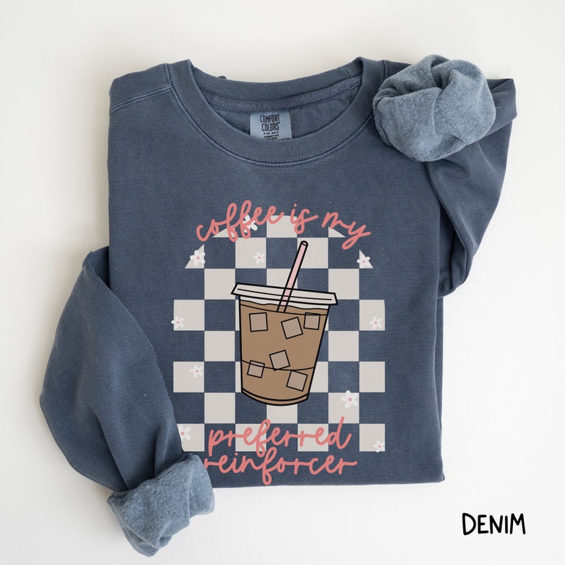 May include: Denim blue sweatshirt with a graphic of an iced coffee drink on a checkered background. The text "coffee is my preferred reinforcer" is in pink script. The sweatshirt has a crew neck and long sleeves.