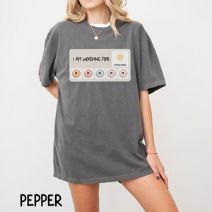 May include: A dark gray t-shirt with a graphic print of a progress bar with smiley faces. The text "I AM WORKING FOR: SUMMER BREAK" is printed above the progress bar.