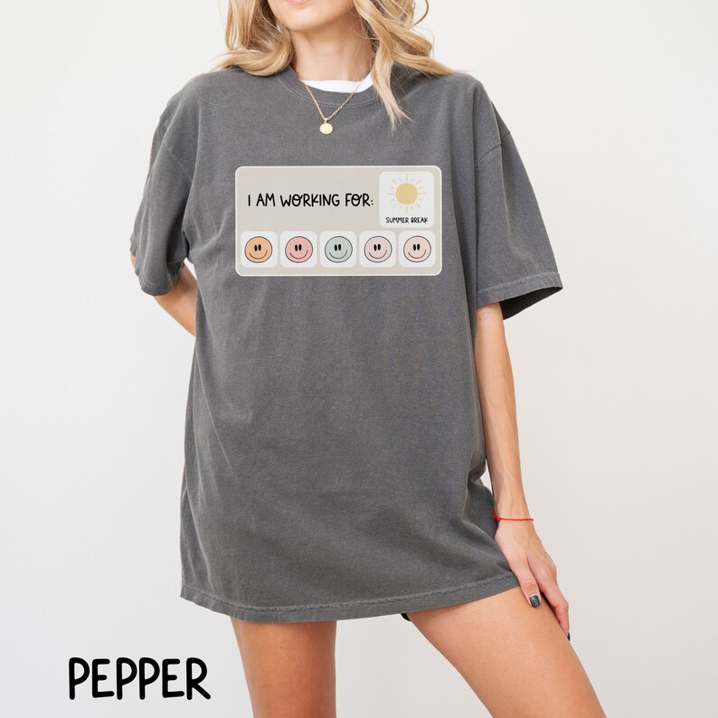 May include: A dark gray t-shirt with a graphic print of a progress bar with smiley faces. The text "I AM WORKING FOR: SUMMER BREAK" is printed above the progress bar.