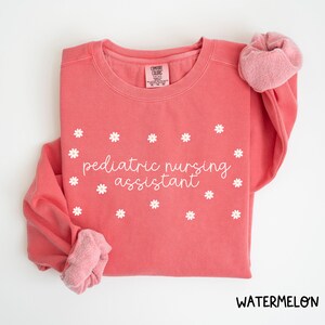 Może przedstawiać: Różowa bluza z napisem "pediatric nursing assistant" w białym kolorze i wzorem białych stokrotek.
