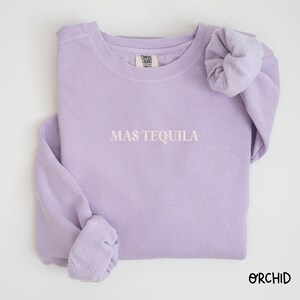 May include: A lavender sweatshirt with the text "MAS TEQUILA" in white embroidery. The sweatshirt is folded and has a scrunchie on one of the sleeves.