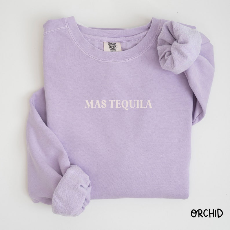 May include: A lavender sweatshirt with the text "MAS TEQUILA" in white embroidery. The sweatshirt is folded and has a scrunchie on one of the sleeves.