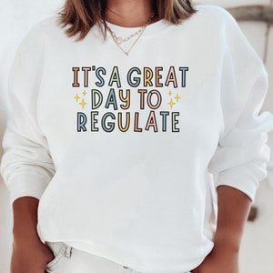 Peut inclure: Sweat-shirt blanc avec le texte "It's a great day to regulate" en lettres color&eacute;es avec des &eacute;toiles jaunes.