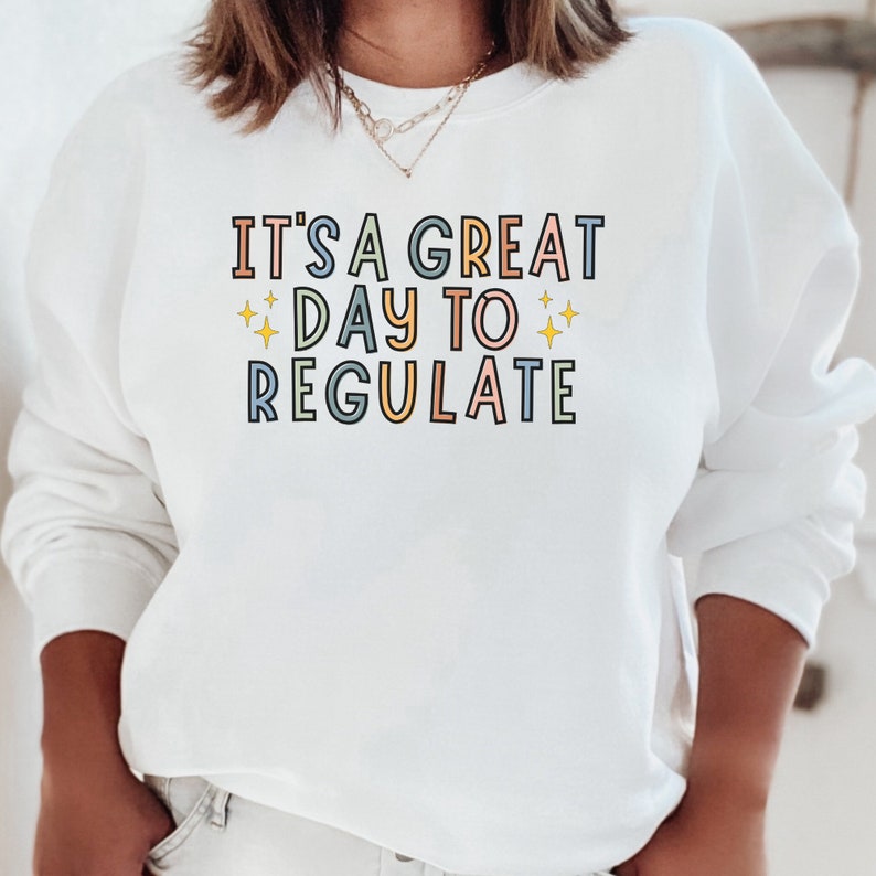 Peut inclure: Sweat-shirt blanc avec le texte "It's a great day to regulate" en lettres color&eacute;es avec des &eacute;toiles jaunes.
