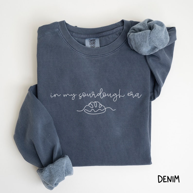 Peut inclure: Un sweat-shirt bleu fonc&eacute; avec le texte "in my sourdough era" et un dessin simple d'une miche de pain. Le sweat-shirt est fait d'un tissu doux et confortable et a une coupe d&eacute;contract&eacute;e.