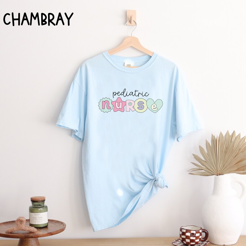 May include: Light blue t-shirt with the text "pediatric nurse" in colorful letters with decorative elements. The letters are outlined in black and have a pastel color scheme.