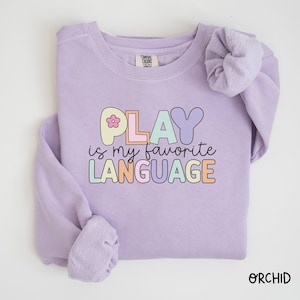 May include: A lavender sweatshirt with the text "PLAY is my favorite LANGUAGE" in colorful letters. The letters are outlined in black and have a flower graphic on the "P".