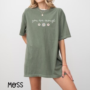 May include: An oversized green t-shirt with the text "you are enough" and a daisy with three smiley faces.