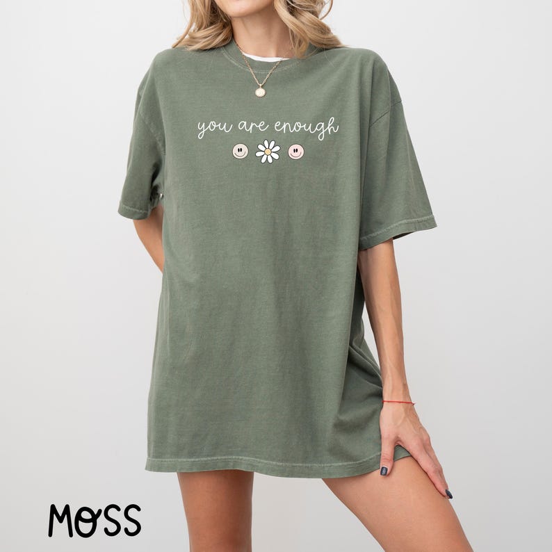 May include: An oversized green t-shirt with the text "you are enough" and a daisy with three smiley faces.