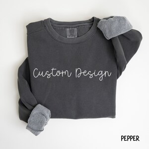 Könnte beinhalten: Ein dunkelgrauer Pullover mit dem Text "Custom Design" in weißen Buchstaben. Der Pullover hat eine Fleece-Futter an der Innenseite der Bündchen.