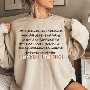 May include: A beige sweatshirt with the text "I'm a scientist practitioner who applies the natural science of behavior to systematically manipulate the environment to improve the lives of others I work with kids".