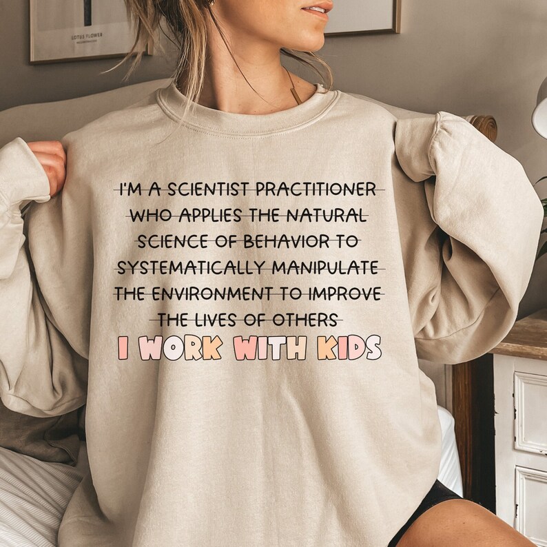 May include: A beige sweatshirt with the text "I'm a scientist practitioner who applies the natural science of behavior to systematically manipulate the environment to improve the lives of others I work with kids".