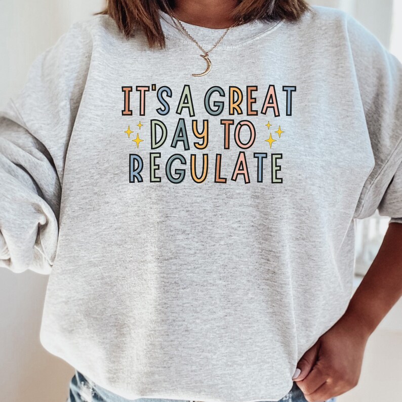 Peut inclure: Sweat-shirt gris avec le texte "It's a great day to regulate" en lettres color&eacute;es avec des &eacute;toiles jaunes de chaque c&ocirc;t&eacute; du mot "day".