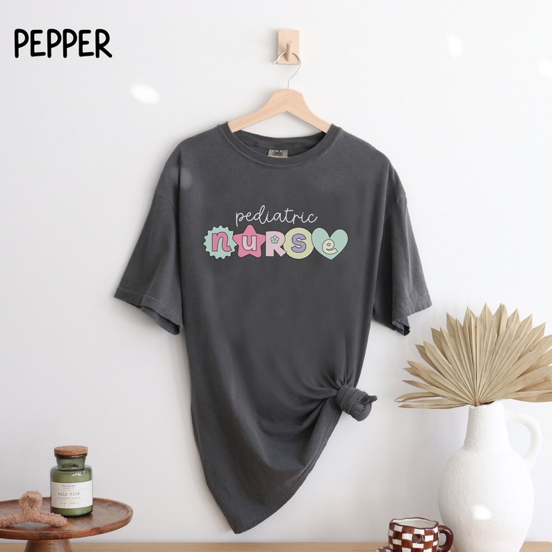 May include: A dark gray t-shirt with the text "pediatric nurse" in colorful letters. The letters are decorated with hearts and stars.
