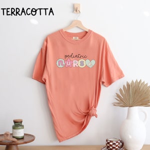 May include: A terracotta colored t-shirt with the text "pediatric nurse" in colorful letters. The letters are decorated with stars and hearts.