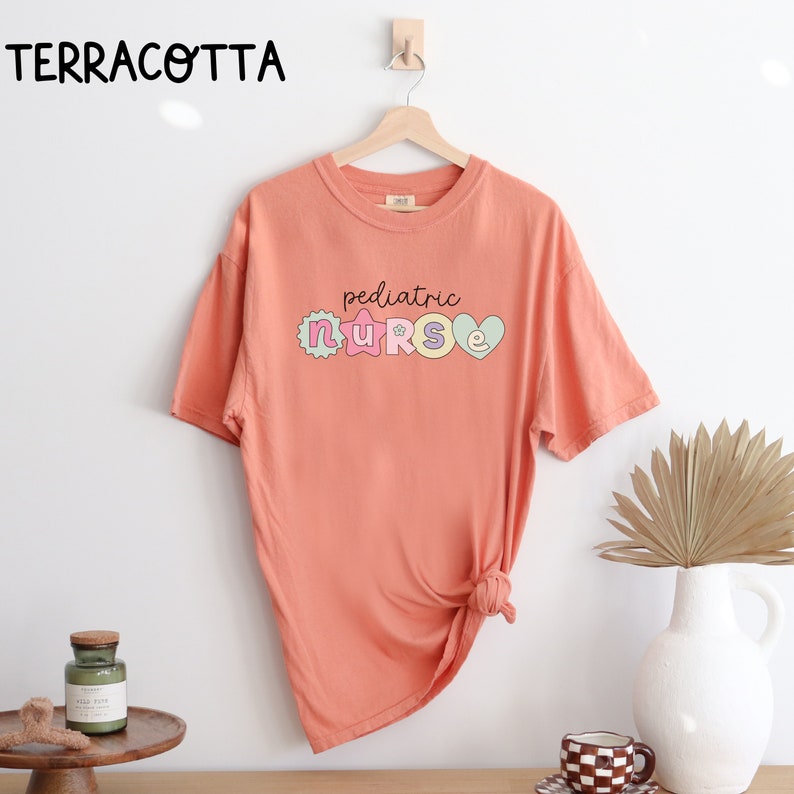 May include: A terracotta colored t-shirt with the text "pediatric nurse" in colorful letters. The letters are decorated with stars and hearts.