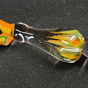 May include: A colorful glass pipe with a swirl design in green, orange, and yellow. The pipe has a clear glass body with a round, iridescent green and blue glass tip.
