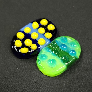 May include: Two oval-shaped glass paperweights with a textured surface. One paperweight is blue, green, and yellow with yellow circles. The other paperweight is green and blue with blue circles.