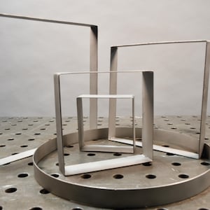 May include: Four metal frames of varying sizes, with the largest frame standing upright and the other three frames nested inside. The frames are all made of a silver-colored metal and have a simple, geometric design. The frames are sitting on a gray, textured surface.