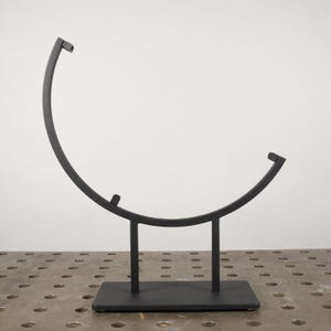 May include: Black metal stand with a curved top, designed to hold a round object. The stand has a rectangular base and three legs.