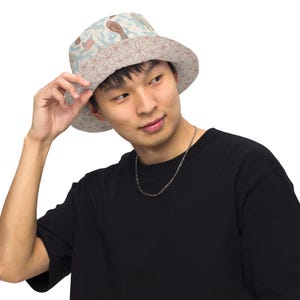 Può includere: Un cappello a secchiello reversibile con sfondo azzurro e motivo floreale marrone e bianco su un lato, e sfondo beige con motivo floreale a contorno marrone sull'altro, caratterizzato da una tesa larga e una corona arrotondata.