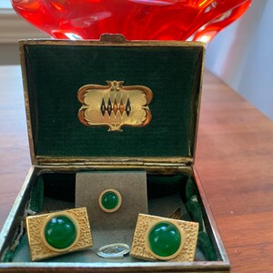 May include: An open vintage jewelry box with a green velvet interior. The box contains a set of gold-toned cufflinks and a matching tie tack, each featuring a large, round, green stone. A clear ring sits in the box.