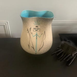 May include: A ceramic pitcher with a light blue rim and a speckled beige glaze. The pitcher features a stylized Native American design in black and blue.