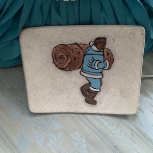 Puede incluir: Un azulejo de cerámica blanco con una ilustración marrón y azul de una persona que lleva un tronco a la espalda. La persona lleva una chaqueta azul y botas marrones.