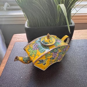 May include: A yellow teapot with a metallic finish featuring a painted pattern of purple grapes, green leaves, and a hummingbird. The teapot has a curved, angular body, a small spout, and a circular lid with a pointed gold-coloured finial on top.