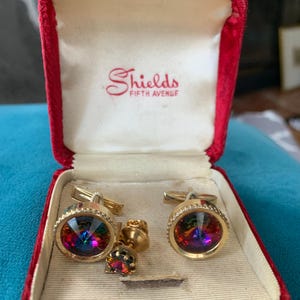 May include: A set of gold-toned cufflinks and a matching shirt stud with iridescent, multi-colored crystal centers, presented in a red velvet box. The box is lined with cream-colored fabric and features the text "Shields FIFTH AVENUE".