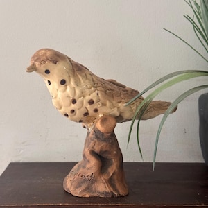 Könnte beinhalten: Eine Keramik-Vogelfigur mit cremefarbenem Körper, braunen Punkten und strukturierter Oberfläche. Der Vogel sitzt auf einem braunen, stumpfartigen Sockel, in dessen Seite eine Signatur eingraviert ist. Die Skulptur zeigt ein detailliertes Federkleid.