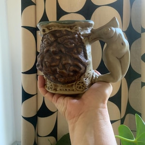 May include: A ceramic mug with a detailed, textured design. The mug features a sculpted face on the front and a nude figure forming the handle. The mug is primarily brown and beige, with a green rim. The mug is held in a hand.