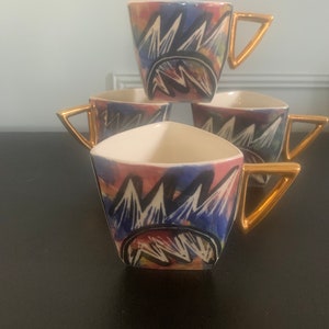 Könnte beinhalten: Vier gestapelte Kaffeetassen mit einzigartiger Form. Jede Tasse hat einen weißen Innenraum, ein farbenfrohes abstraktes Design mit Blau-, Rot- und Gelbtönen und einen goldfarbenen dreieckigen Griff. Die Tassen sind auf einer dunklen Oberfläche angeordnet.
