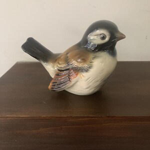 May include: A ceramic bird figurine with a glossy finish. The bird has a black head, a brown and tan body, and a white belly. The figurine is perched on a dark wooden surface.