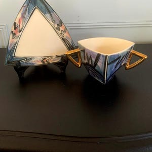 May include: A triangular ceramic teacup and saucer set with abstract geometric designs in shades of blue, purple, and pink. The cup has gold handles and the saucer has a white centre.