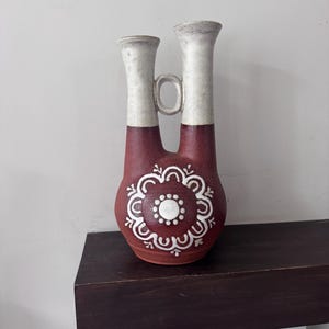 May include: A dual-spout ceramic vase featuring a deep reddish-brown base with a white floral pattern and a solid white upper section. The two necks are connected by a small circular handle, creating a distinct silhouette with a matte finish.