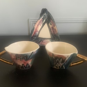 May include: Two ceramic teacups with gold handles and abstract black, blue, pink, and white designs. A triangular ceramic saucer with a similar design.