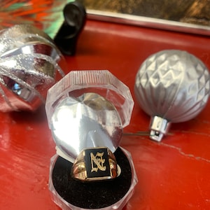 Peut inclure: Une bague chevalière dorée avec une face rectangulaire noire, ornée d'un motif initiales. La bague est présentée dans un écrin transparent articulé sur une surface rouge, avec des boules de Noël argentées en arrière-plan.