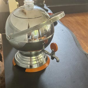 May include: A spherical chrome beverage dispenser with a polished reflective surface, a clear glass knob on the lid, and a prominent orange handle on the spigot. The base features a matching orange circular trim, and the unit has a swing-style handle.