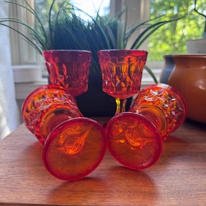 May include: A collection of five vintage orange-red glass cordial glasses. The glasses have a textured diamond pattern on the bowl and a delicate stem. The glasses are arranged on a wooden surface, with a plant in the background.