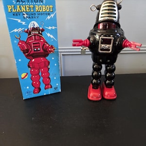 Può includere: Un robot giocattolo vintage e la sua scatola. Il robot è nero con dettagli rossi, tra cui mani e piedi. La scatola è blu con le parole "ACTION PLANET ROBOT" e un'illustrazione di robot rosso. Il robot è alto circa 20 cm.